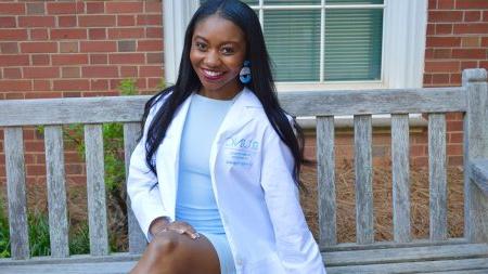 Brooklyn Rushing sitting on a bench wearing a Carolina Blue dress and a white lab coat.