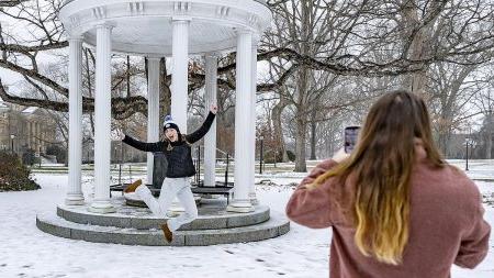 A student standing in the foreground taking a photo of her friend leaping in front the Old Well during the winter season.